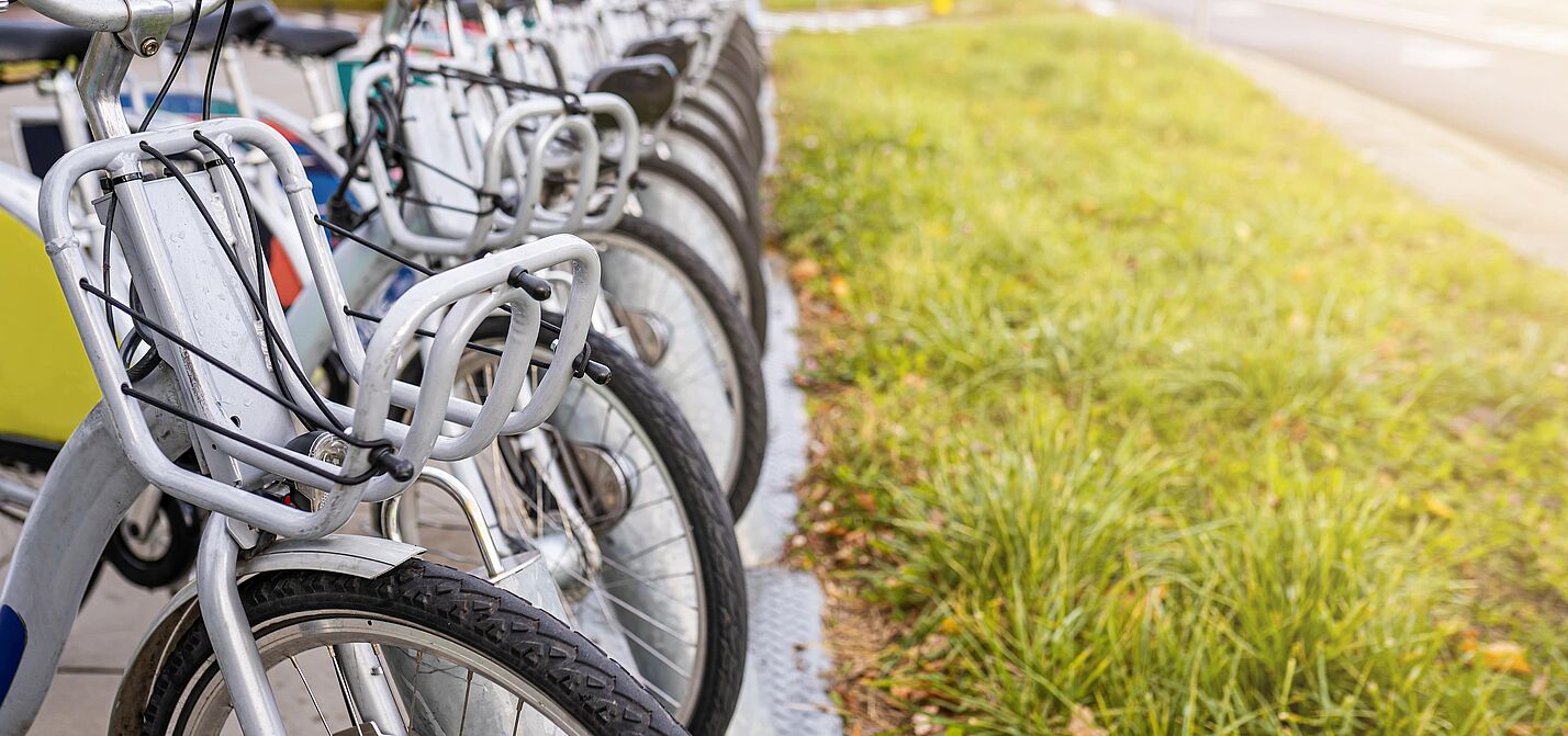 Öffentliche Leihfahrräder an einer Bikesharing-Station Eine Reihe von öffentlichen Leihfahrrädern mit silbernen Rahmen und grauen Körben am Lenker steht ordentlich nebeneinander an einer stationsgebundenen Verleihstation. Die Fahrräder sind auf einem gepflasterten Gehweg abgestellt, neben dem eine Grünfläche verläuft.